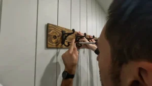 Finished rustic coat rack with six black cast iron hooks mounted on a stained wood plank, installed on a shiplap wall in a mudroom.