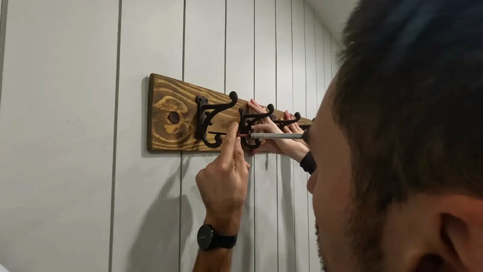 Finished rustic coat rack with six black cast iron hooks mounted on a stained wood plank, installed on a shiplap wall in a mudroom.