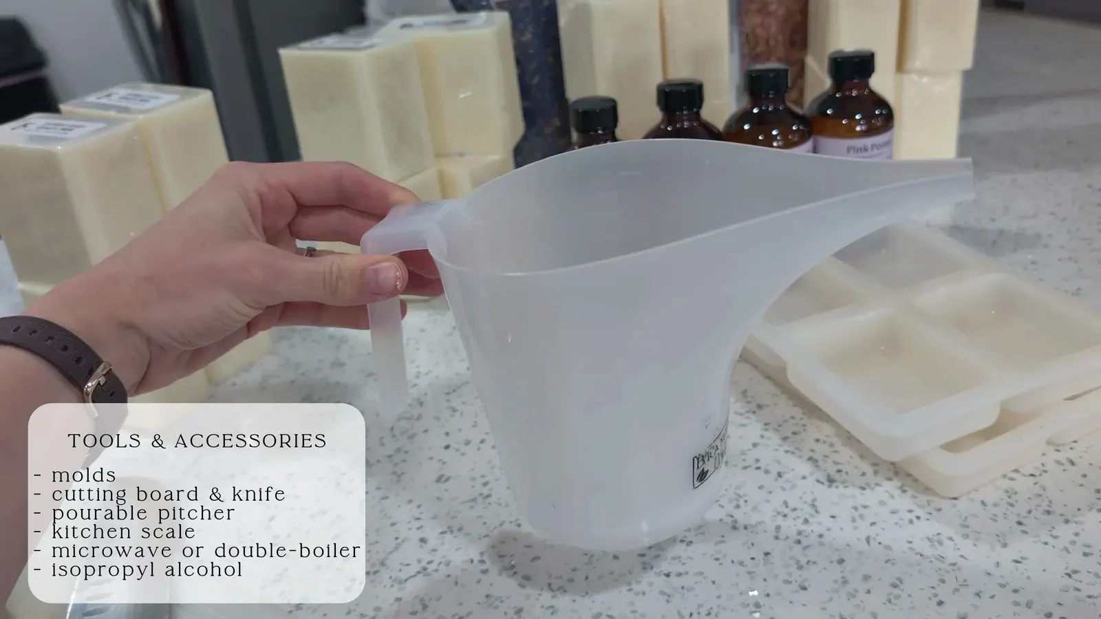 Essential soap making tools arranged on a counter, including silicone molds, a cutting board, a pourable pitcher, a digital scale, and a spray bottle.