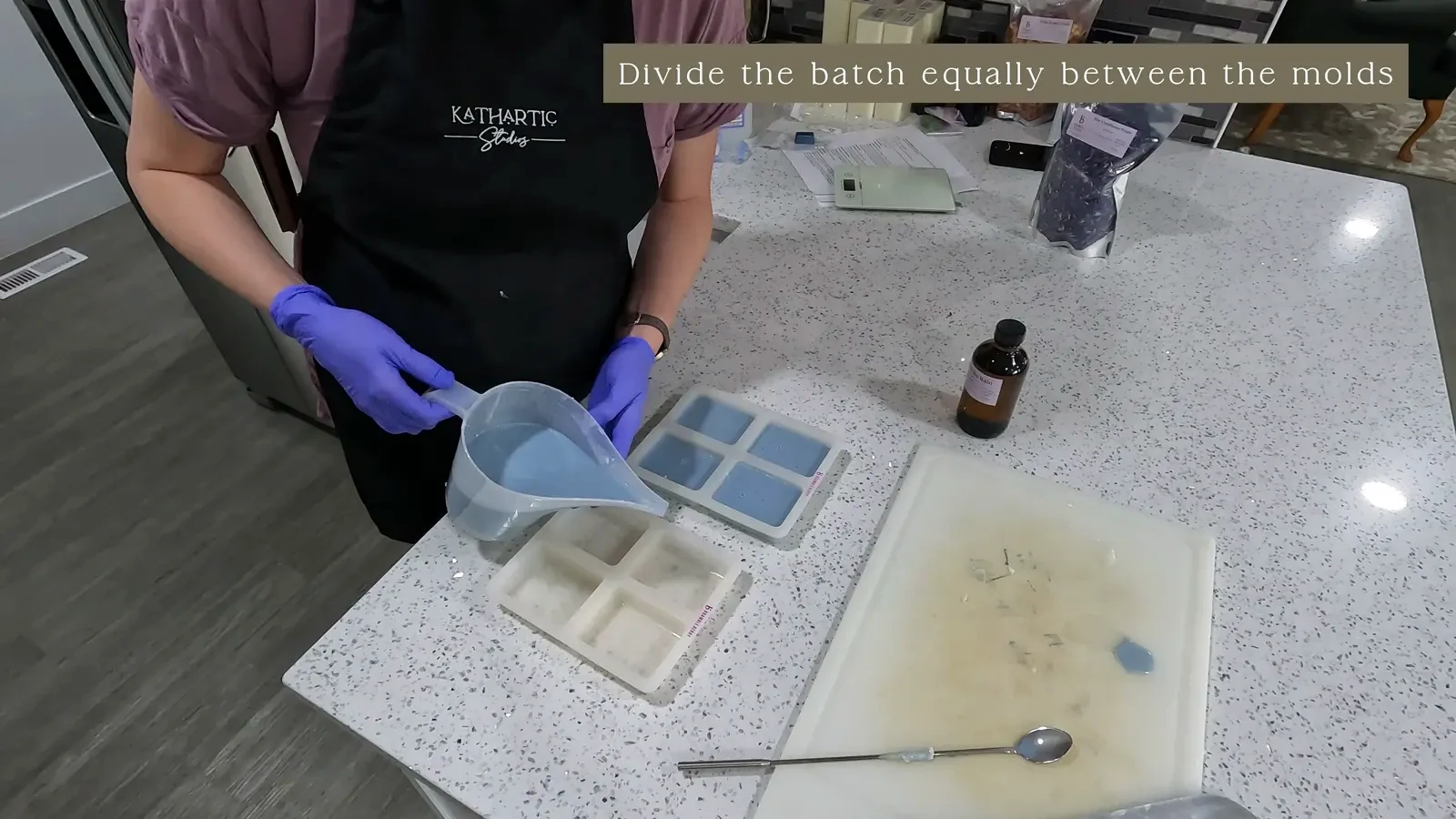 Melted blue soap being poured from a pitcher into multiple silicone molds, with some bubbles visible on the surface.