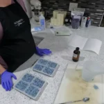 A hand gently pressing on the bottom of a silicone mold to release a finished, blue, flower-shaped soap bar onto a paper towel.