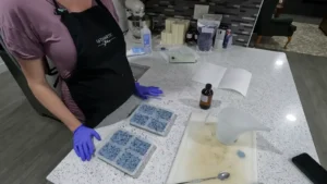 A hand gently pressing on the bottom of a silicone mold to release a finished, blue, flower-shaped soap bar onto a paper towel.