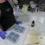 A hand gently pressing on the bottom of a silicone mold to release a finished, blue, flower-shaped soap bar onto a paper towel.