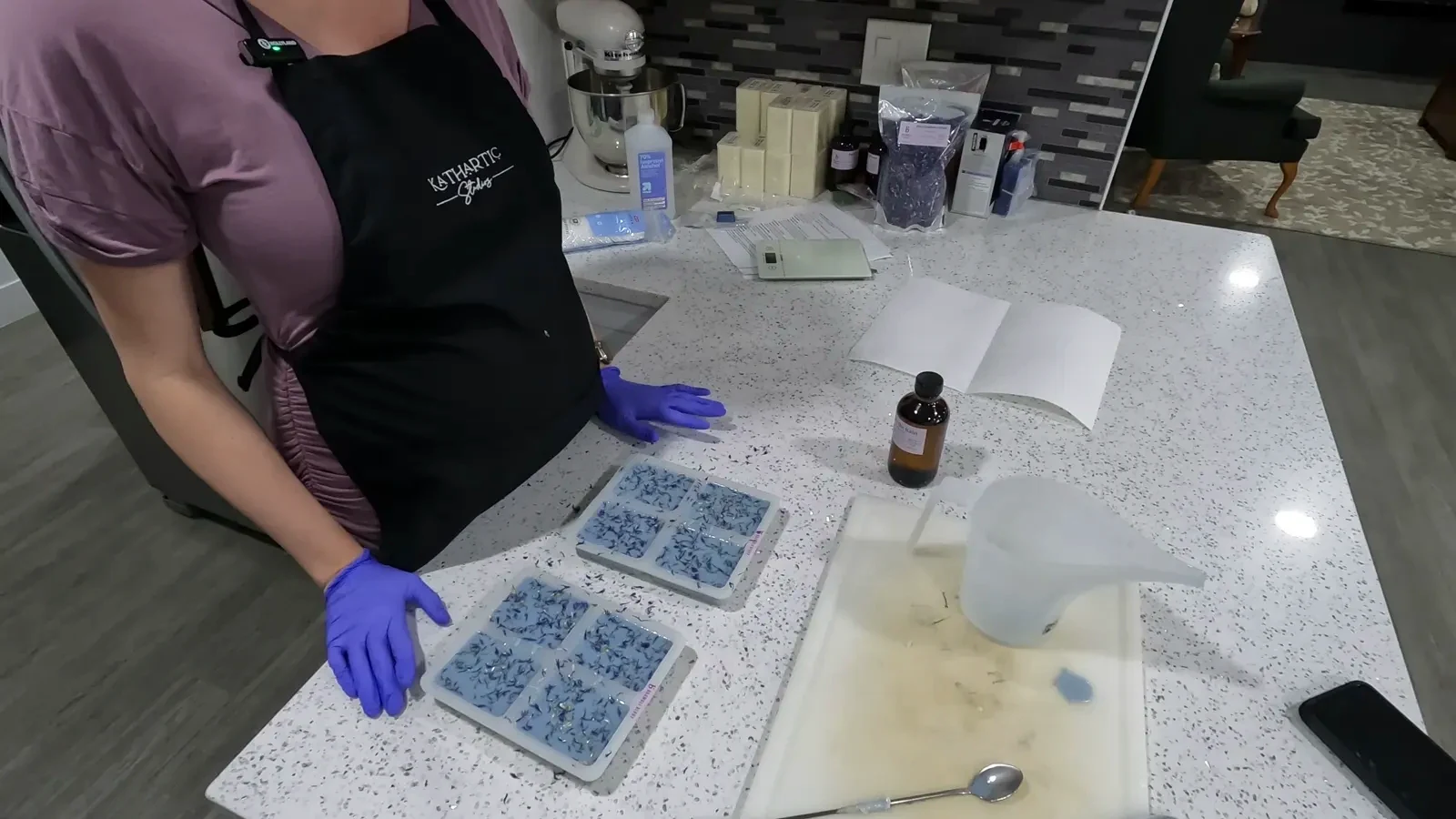 A hand gently pressing on the bottom of a silicone mold to release a finished, blue, flower-shaped soap bar onto a paper towel.