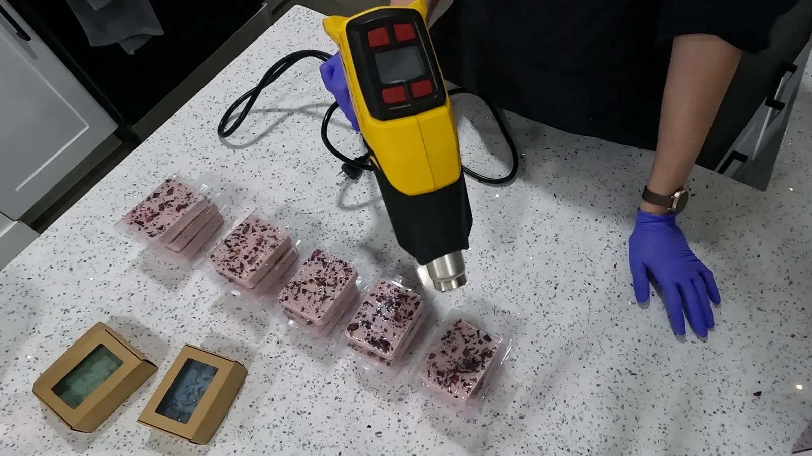 A heat gun being used to shrink a clear plastic wrap tightly around a bar of soap, creating a smooth, sealed package.