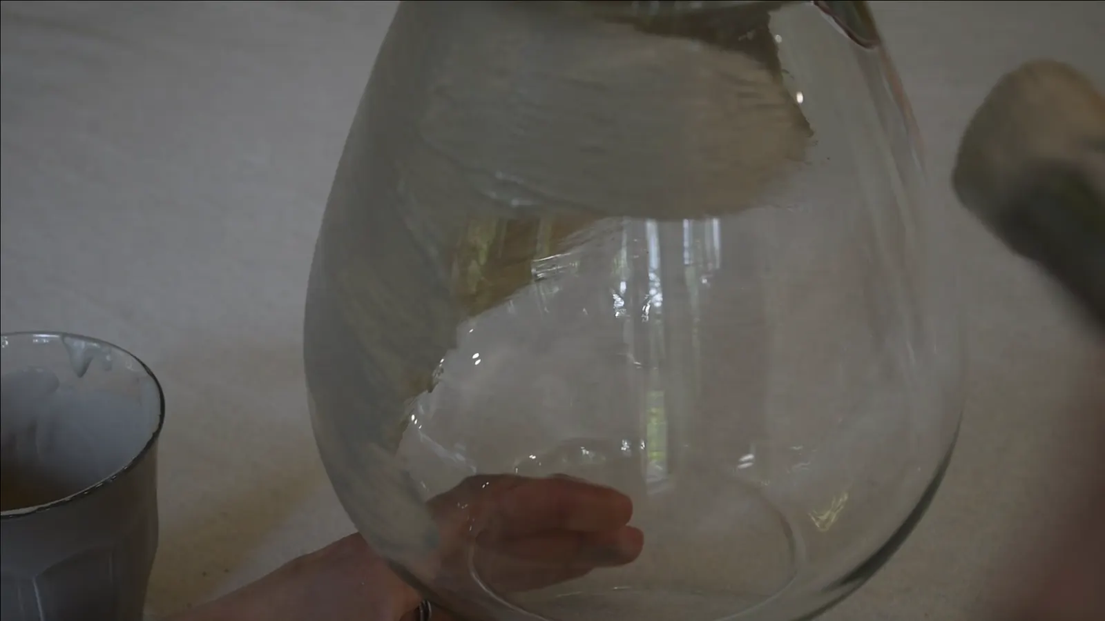 A hand applying the textured beige paint to a glass vase with horizontal brush strokes.