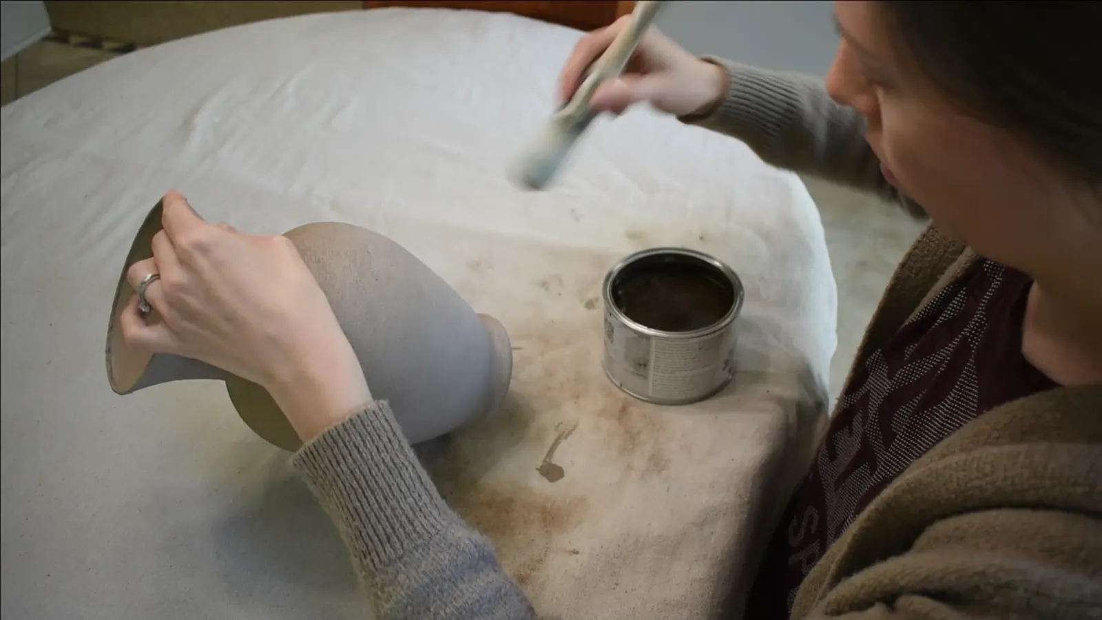 A hand applying brown furniture wax to the textured beige vase with a round paintbrush.