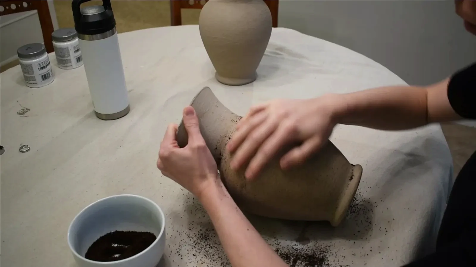 Hands rubbing coffee grounds onto the textured surface of a painted vase to add an aged effect.